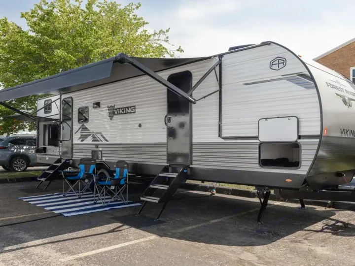 This white Viking travel trailer, a spacious rv rental, with extended awning, two camping chairs, and striped mat for outdoor comfort.. Forest River Viking 6k 292TB 2025