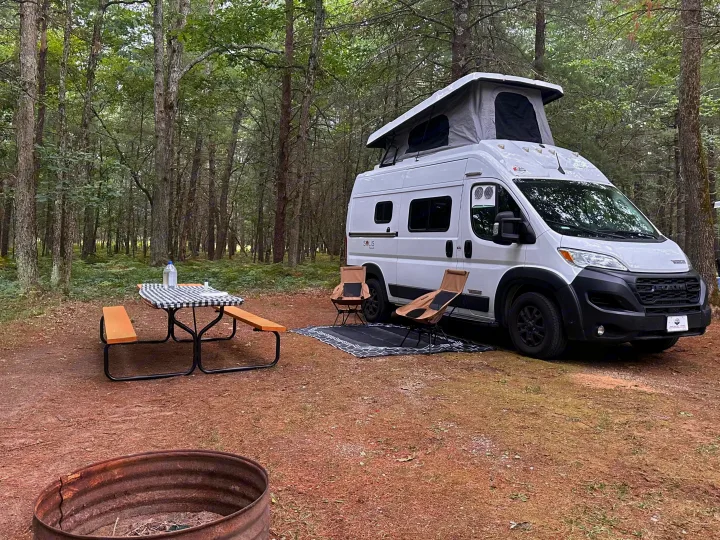 A serene rv rental campsite featuring a white camper van with a pop-top, picnic table, chairs, and a fire pit amidst trees.. Winnebago Solis 2025