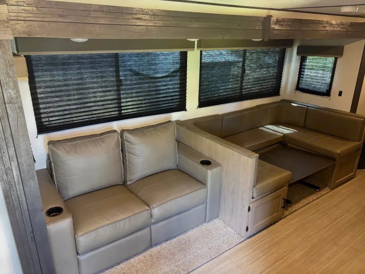 RV rental interior: light tan U-shaped dinette & sofa with cup holders, dark window blinds, and wood-look trim.. Keystone RV Hideout 2021