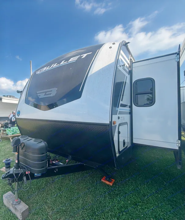 A white and black Bullet travel trailer with an open side door and propane tanks, parked on grass. Ideal for an rv rental.. Keystone RV Bullet Premium 2025