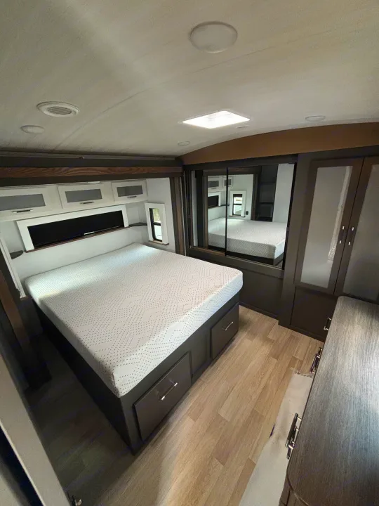 RV rental bedroom showcasing a bed with drawers, mirrored closet doors, and overhead storage. A compact and functional space.. Keystone RV Bullet Premium 2025