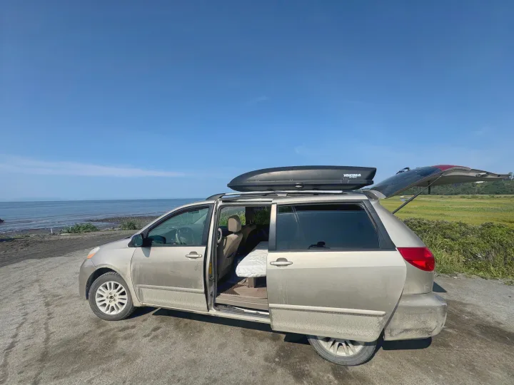 Minivan campered up like an RV rental, doors open to reveal a bed, parked by the ocean. Roof rack has a large cargo box.. Toyota Sienna 2009