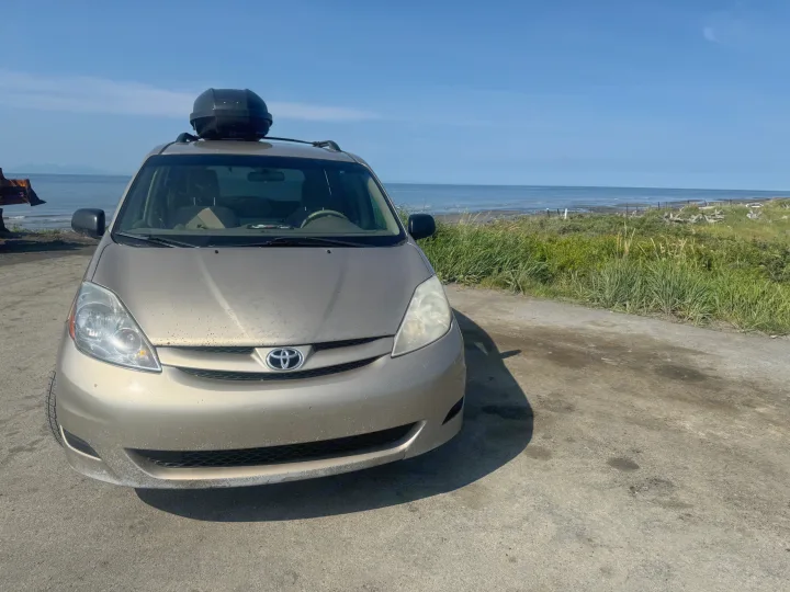 This gold minivan RV rental with a roof box is parked by a serene ocean under a clear blue sky, ideal for coastal camping.. Toyota Sienna 2009