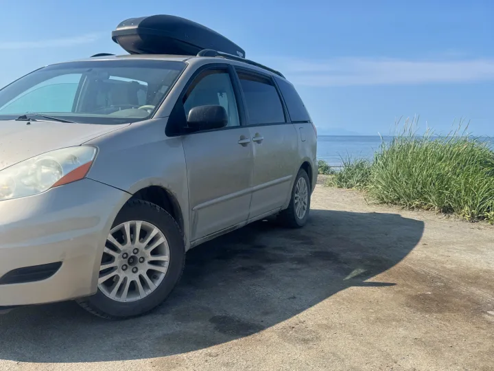 Dusty minivan with roof box by the ocean, next to tall grass. Ideal for coastal road trips, offering adventure akin to an RV rental.. Toyota Sienna 2009