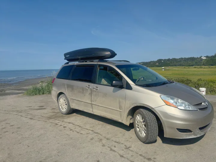A dirty gold minivan with roof box by the ocean, ready for a road trip. A practical travel vehicle, often chosen over an RV rental.. Toyota Sienna 2009