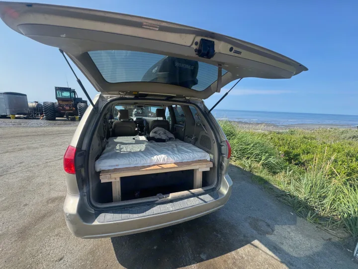 This car, used as an rv rental, has its trunk open, showing a built-in bed by the ocean. A tractor is nearby.. Toyota Sienna 2009