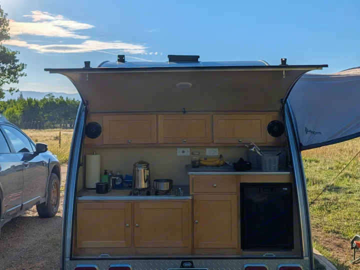 Open galley kitchen of a teardrop RV rental, showing cabinets, sink, stove, and mini-fridge amidst nature.. nuCamp T@B CS-S 2017