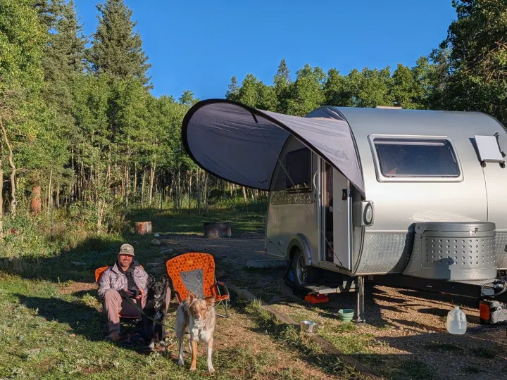 Sunny rv rental camping scene: man and two dogs relaxing beside a silver teardrop trailer with an extended awning in a forest.. nuCamp T@B CS-S 2017