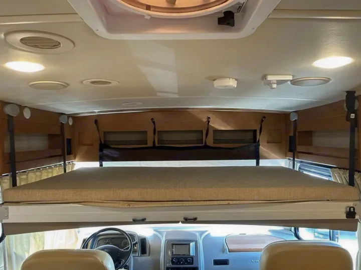 Inside an RV rental, showing the over-cab bunk bed with mattress above the driver's area. Ceiling lights & vents visible.. Winnebago Vista LX 2017