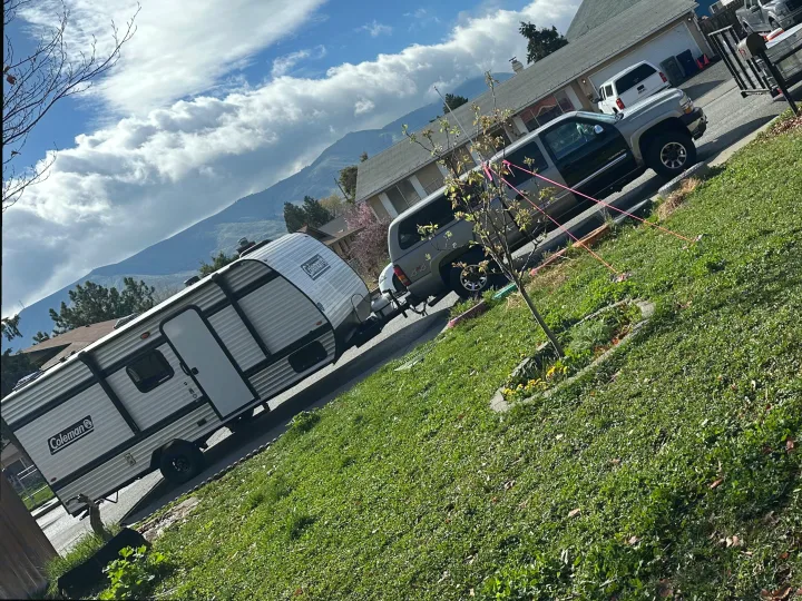 A white & gray Coleman RV rental, hitched to a silver SUV on a street with mountains & cloudy sky in background, ready for travel.. Coleman 17B 2025