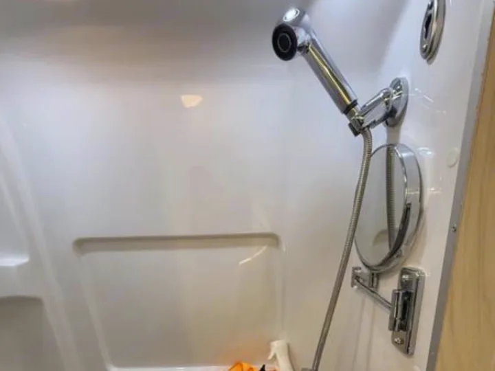 RV rental shower with a chrome handheld showerhead, hose, and a pivoting wall-mounted mirror in a white stall.. Airstream Interstate 2025