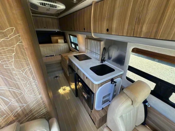 RV rental interior showcasing a modern kitchen with sink/stovetop, wood cabinets, and light-colored seating in a compact camper van.. Airstream Interstate 2025