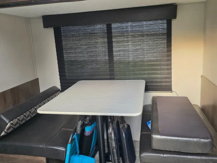 An RV rental dinette featuring a light table, dark upholstered benches, and a window with drawn pleated privacy blinds.. Avenger LT 2021