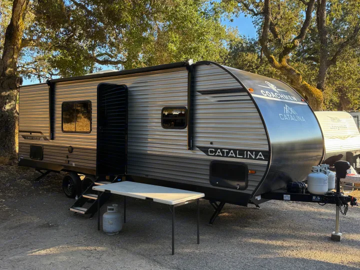 A gray and black Coachmen Catalina RV rental is parked outdoors under trees, with an open door, folding table, and propane tanks.. Coachmen Catalina 261 bh 2026