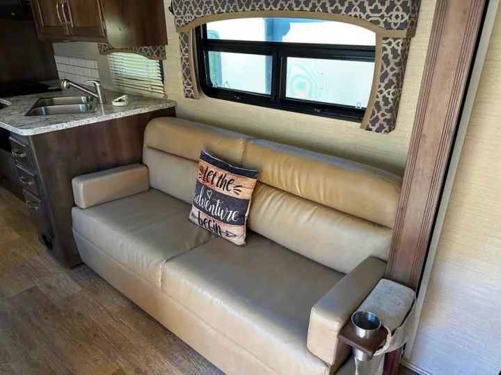 Cozy RV rental interior shows a light-colored sofa with an "adventure" pillow, facing a window and partial kitchen.. Jayco Alante 2018