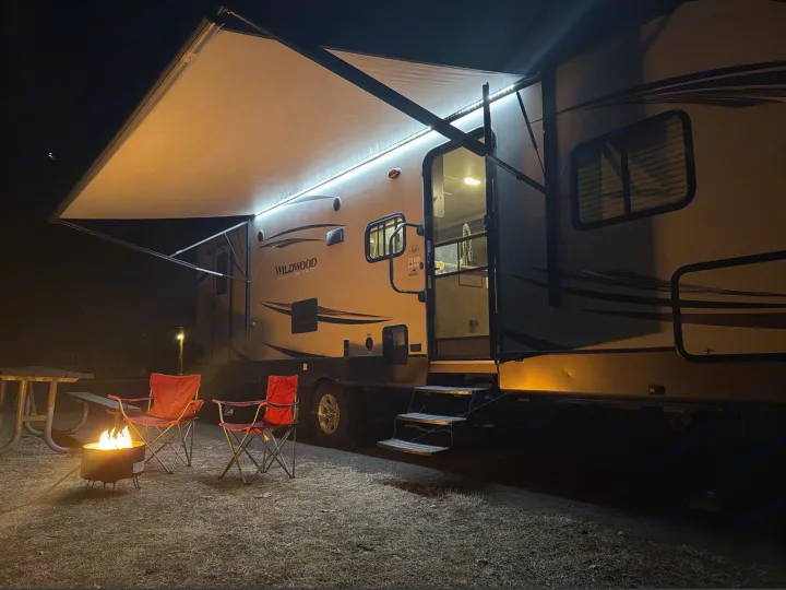 An RV rental illuminates a night campsite. Its awning glows over a burning fire pit and two red camping chairs.. Forest River T27TDSS 2017