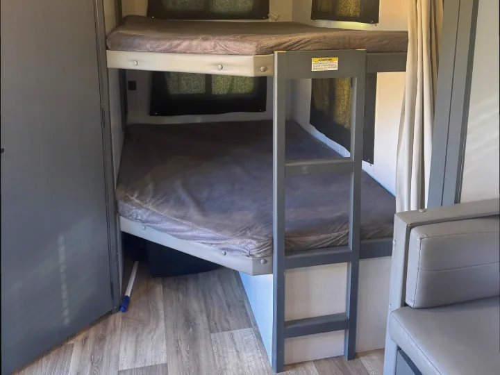 Inside an rv rental, spacious bunk beds with grey bedding, dark window blinds, and a built-in ladder for comfortable sleep.. Keystone RV Bullet Ultra Lite 2022