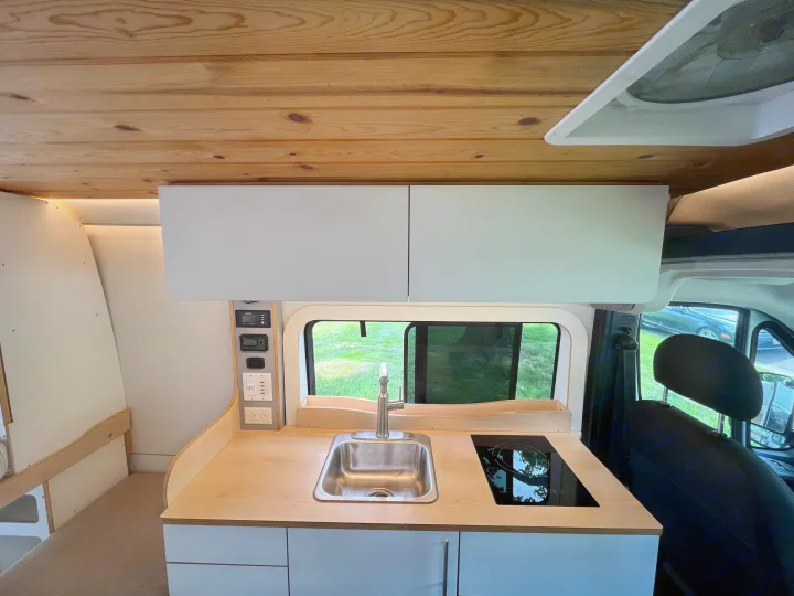 Modern rv rental van kitchen: wood ceiling, light gray cabinets, counter with sink, induction cooktop, and window view.. Dave & Matt Vans Dave & Matt Vans Class B 2023