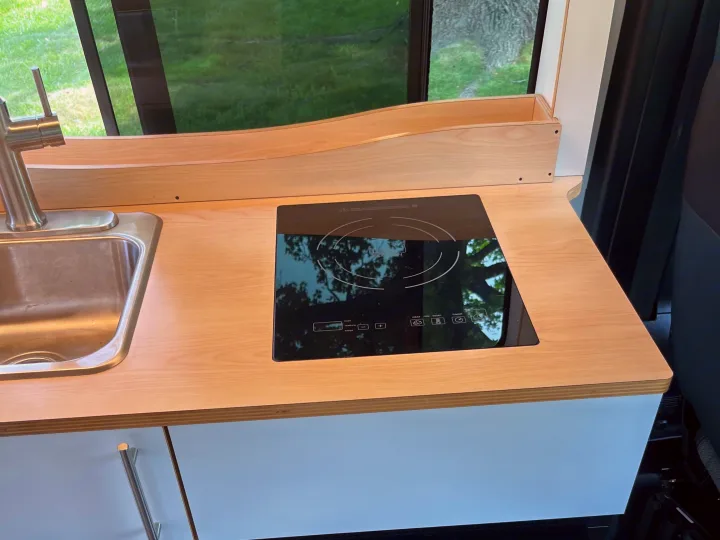 Modern RV rental kitchen with integrated induction cooktop, stainless steel sink, and light wood countertop by a window.. Dave & Matt Vans Dave & Matt Vans Class B 2023