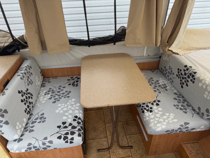 RV rental pop-up camper dinette with grey leaf-patterned cushions, a light brown table, and a screened window.. Forest River Rockwood Freedom 2013