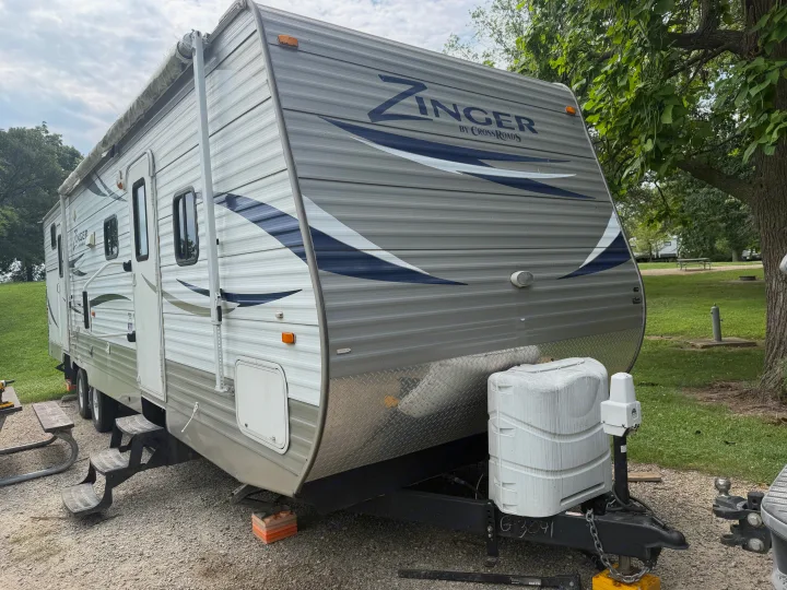 Zinger travel trailer rv rental parked at a campsite. The grey RV shows its front, side, steps, and rolled-up awning.. Crossroads RV Zinger 2012