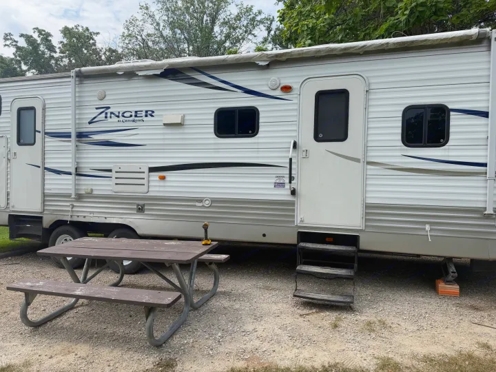 White Zinger travel trailer rv rental, complete with a rolled awning and steps, parked at a gravel campsite with a picnic table.. Crossroads RV Zinger 2012