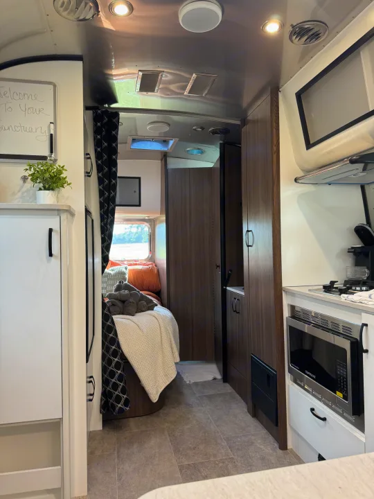 Modern rv rental interior, featuring a cozy bedroom, sleek kitchen appliances, and a welcoming 'Welcome To Your Sanctuary' sign.. Airstream Caravel 2023