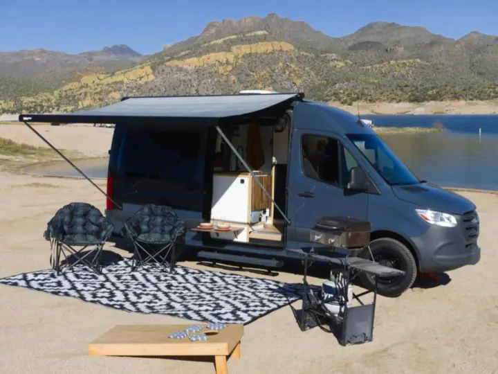 RV rental camper van parked by a lake with mountains, awning extended, and outdoor camping setup with chairs.. Mercedes Sprinter 2500 2019