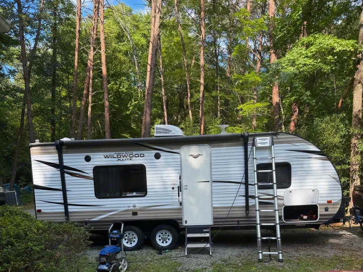 A white Forest River Wildwood X-Lite RV rental is parked among tall trees. A ladder and outdoor gear are visible.. Forest River Salem Cruise Lite 2018