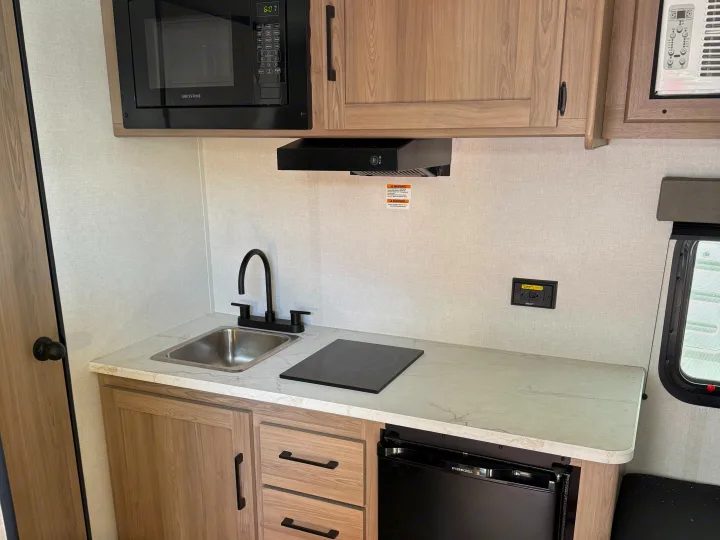 Step into this bright RV rental kitchen featuring a Greystone microwave, sleek sink, induction cooktop, and EverChill fridge.. Keystone RV Coleman 2026