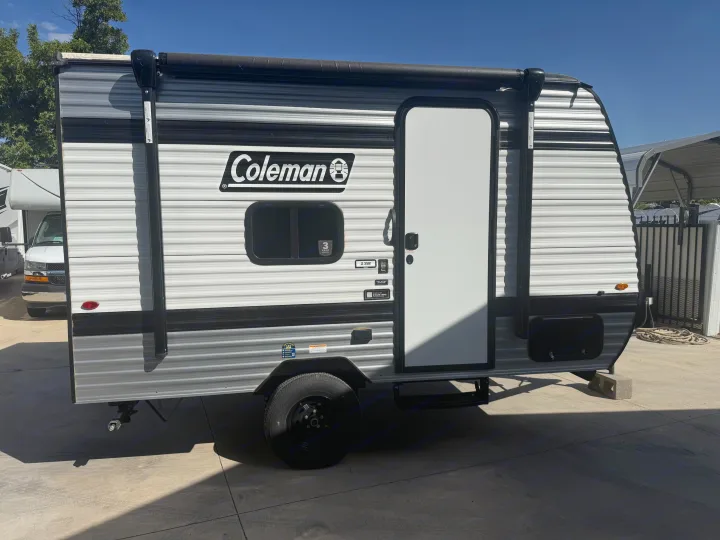 Sunny day view of a Coleman travel trailer rv rental, featuring gray/white striped siding, black door, window, and rolled awning.. Keystone RV Coleman 2026