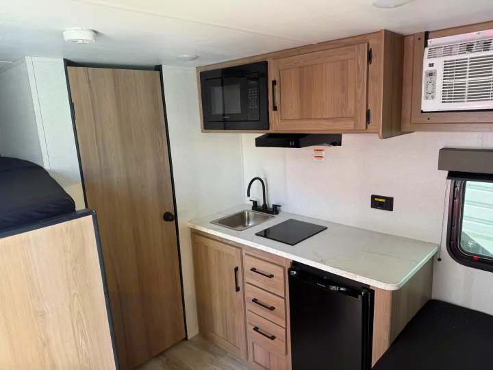 Modern RV rental interior: kitchenette with wood-look cabinets, microwave, fridge, sink, and cooktop. AC unit above.. Keystone RV Coleman 2026