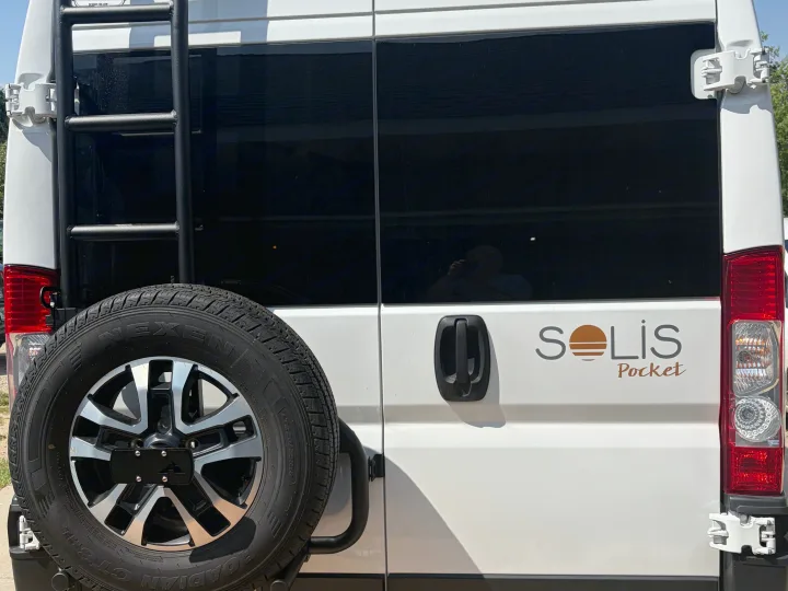 Rear view of a white Solis Pocket rv rental, featuring a black ladder, spare tire, and dark tinted windows.. Winnebago Solis 2025