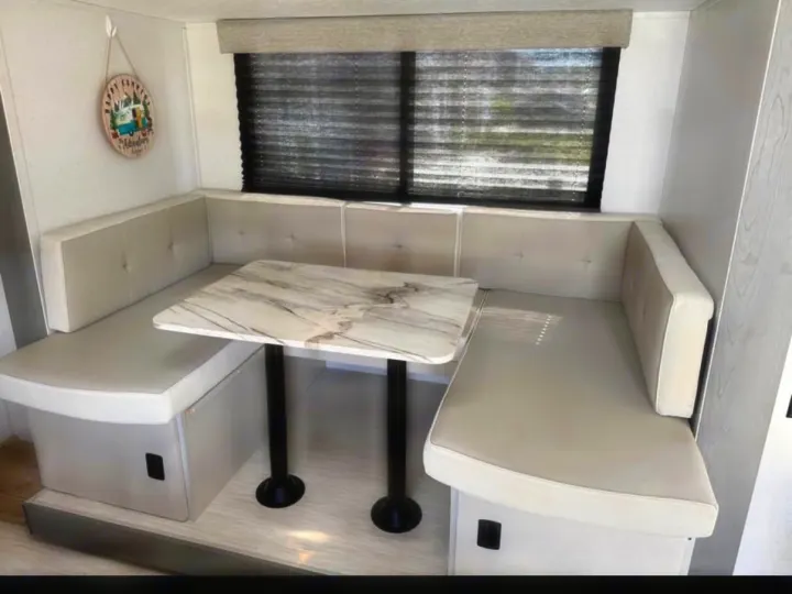 This RV rental features a modern dinette with light beige U-shaped seating, a marble-look table, and a window with black blinds.. Forest River Wildwood FSX 2023