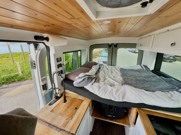 Inside this **rv rental**: a cozy bed, wood ceiling, and compact kitchen with a sink near an open door, ready for adventures.. Ford Transit 2018