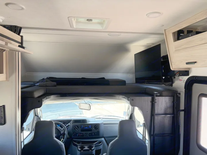 This RV rental interior features an over-cab bunk bed, wall-mounted TV, and front vehicle dashboard.. Forest River Sunseeker LE 2024