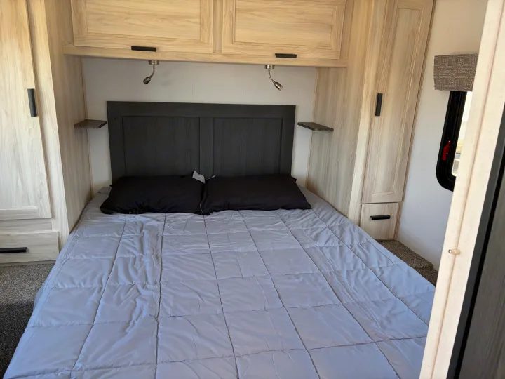 Cozy RV rental bedroom featuring a full bed with a gray comforter, dark headboard, light wood cabinetry, reading lights, and side storage.. Forest River Sunseeker LE 2024