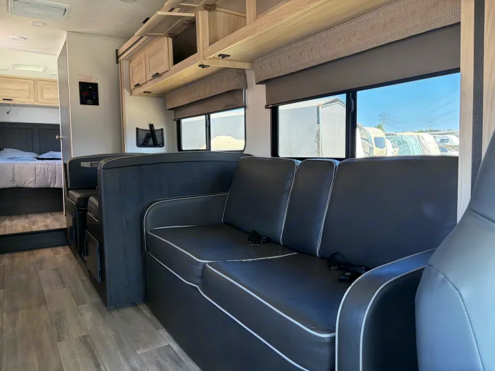 Bright rv rental interior featuring a cozy dark grey seating area with white piping, overhead cabinets, and a rear bedroom.. Forest River Sunseeker LE 2024