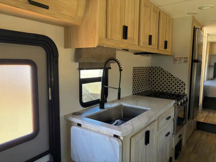 Spacious RV rental kitchen with modern light wood cabinets, marble-look counters, extendable flap, sink, stove, and fridge.. Forest River Sunseeker LE 2024
