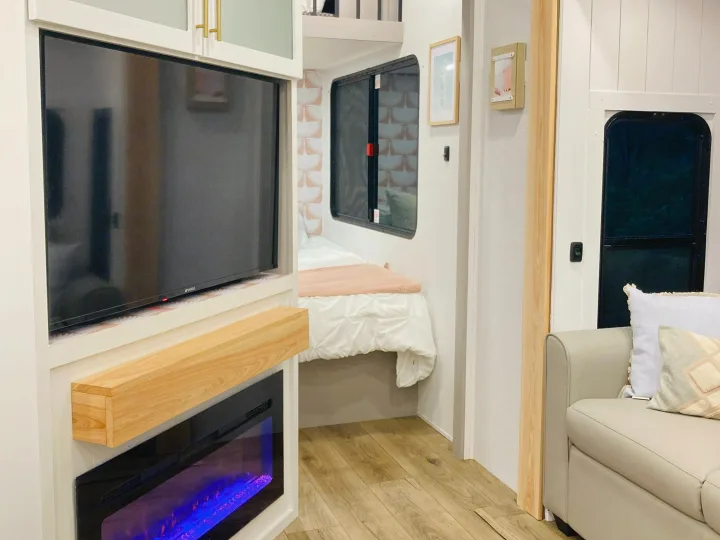 Modern RV rental interior with a TV, electric fireplace, bed, and sofa, accented by light wood and white finishes.. Alliance RV Avenue 2026