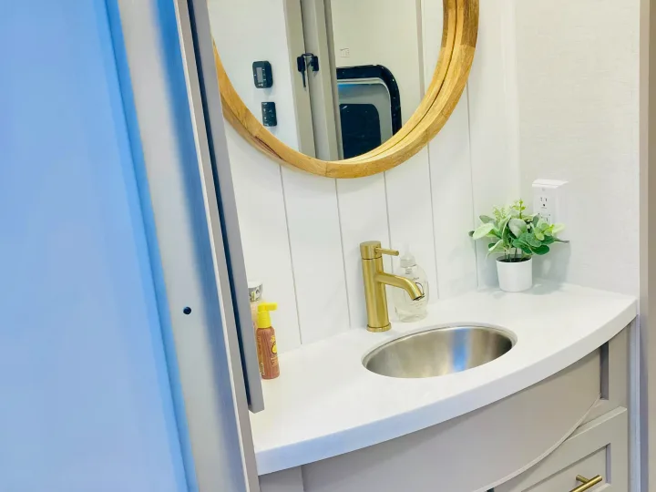 Stylish RV rental bathroom featuring a gold faucet, round mirror, white vanity, and shiplap wall. Bright and functional.. Alliance RV Avenue 2026