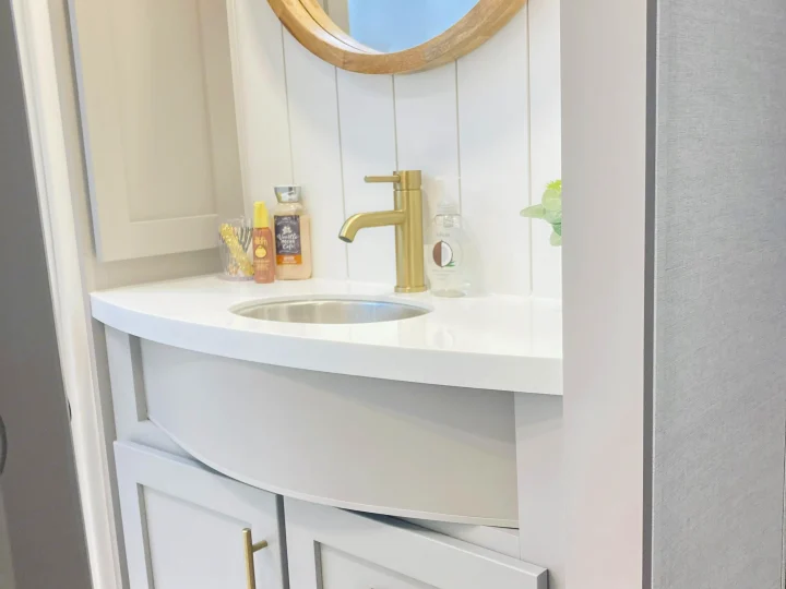 Modern RV rental bathroom vanity. White countertop, gold faucet, round mirror, and light gray cabinets with gold pulls.. Alliance RV Avenue 2026