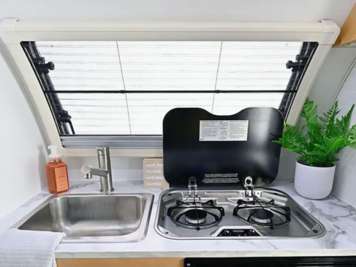Cozy RV rental kitchen: stainless steel sink, 2-burner stove with cover, soap, and plant under a bright window.. nuCamp T@B 2026