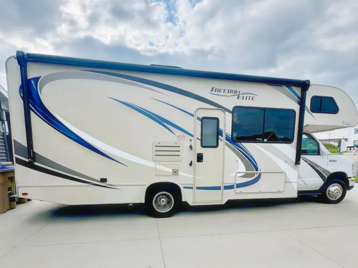 A beige "Freedom Elite" RV rental with blue and gray graphics is parked on a paved surface under a cloudy sky.. Thor Freedom Elite 2017