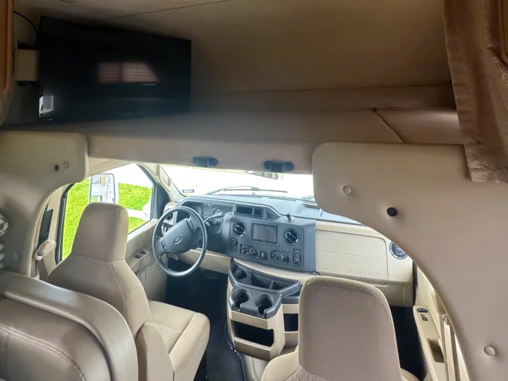 RV rental interior: light beige cockpit with dashboard, steering wheel, front seats, and an overhead TV screen mounted above.. Thor Freedom Elite 2017