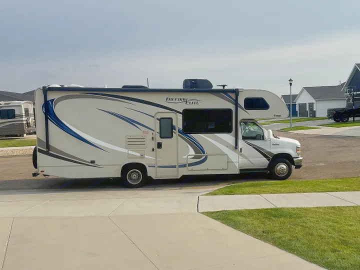 A beige and blue Thor Freedom Elite RV, possibly an RV rental, is parked. This motorhome with an awning is ready for your next adventure.. Thor Freedom Elite 2017