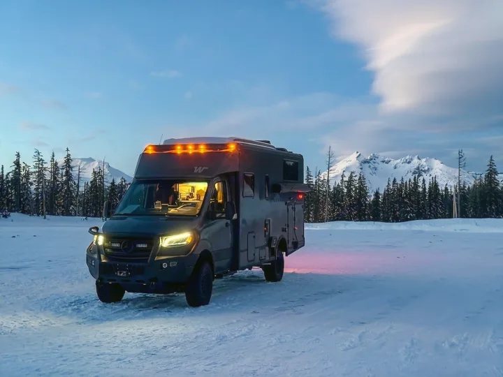 Rugged gray RV rental parked in a vast snowy field. Its lights glow, casting warmth against the twilight, snow-capped mountains, and pines.. Winnebago Ekko 2025