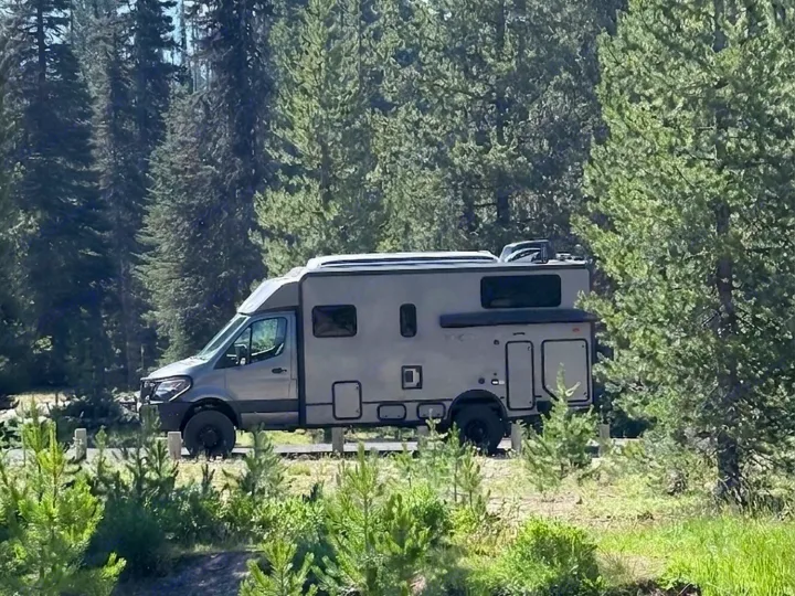 Rugged grey RV rental camper van parked in a sunlit forest, ready for adventure among towering pine trees.. Winnebago Ekko 2025