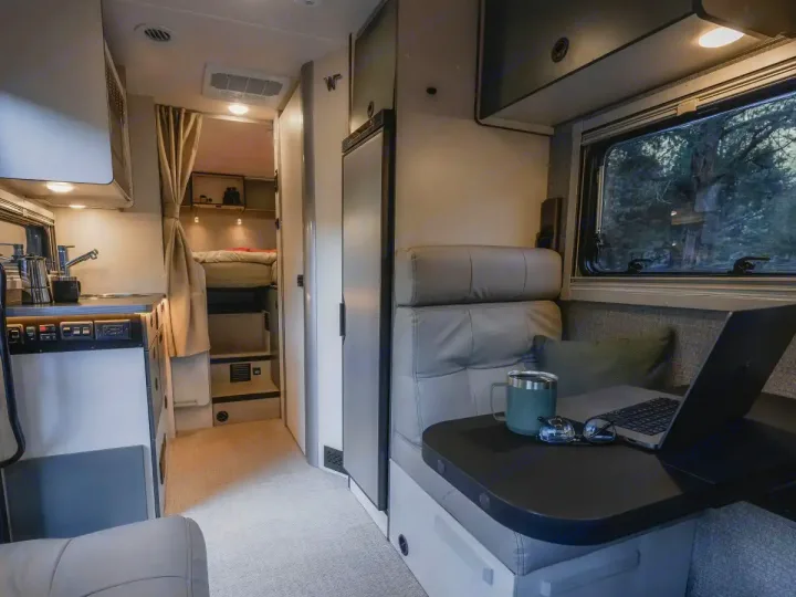 Modern RV rental interior showing a compact kitchen, workspace with laptop, comfy seating, and rear sleeping quarters.. Winnebago Ekko 2025