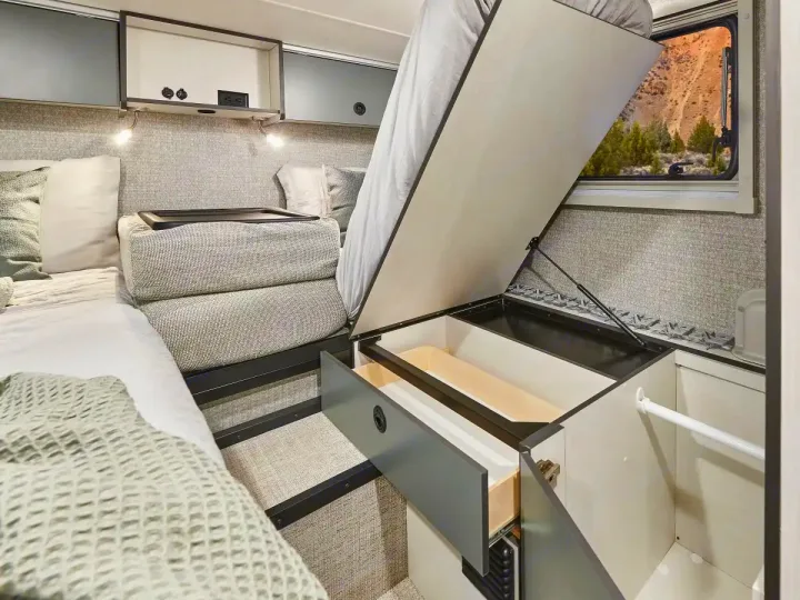 This modern RV rental's interior shows a bed lifted for under-bed storage, open drawers, overhead bins, and a scenic mountain window view.. Winnebago Ekko 2025
